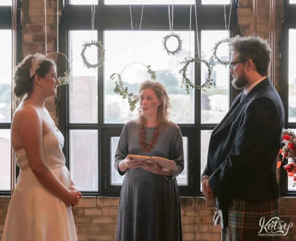 An officient wearing a red necklace and holding a notebook speaks to a bride and groom during their Burroughes Building wedding ceremony in Toronto, Canada