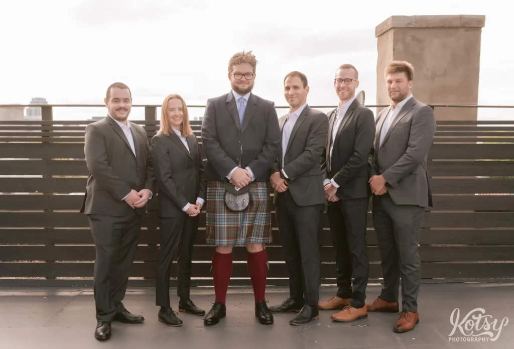 A groom wearing a gray blazer, blue kilt and red stockings poses with his groomsmen on a rooftop at The Burroughes Building in Toronto, Canada