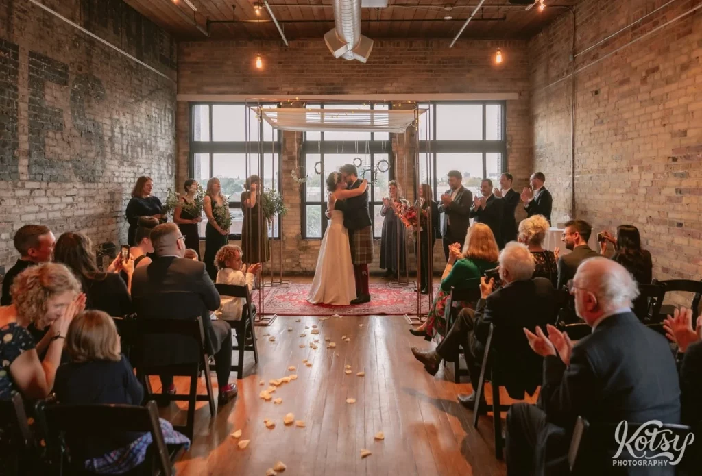 Wide shot of a bride and groom kissing as their wedding party and guests look on during their Burroughes Building wedding ceremony in Toronto, Canada