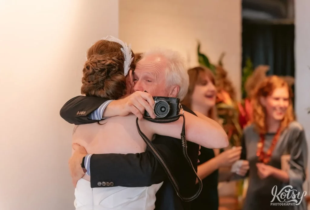 A man holding a small camera hugs his daughter after her Burroughes Building wedding ceremony in Toronto, Canada