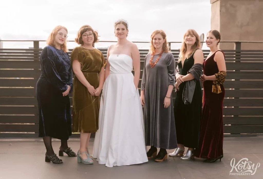 A bride and a white wedding gown poses with her bridesmaids wearing different colored dresses on the rooftop patio at The Burroughes Building in Toronto, Canada