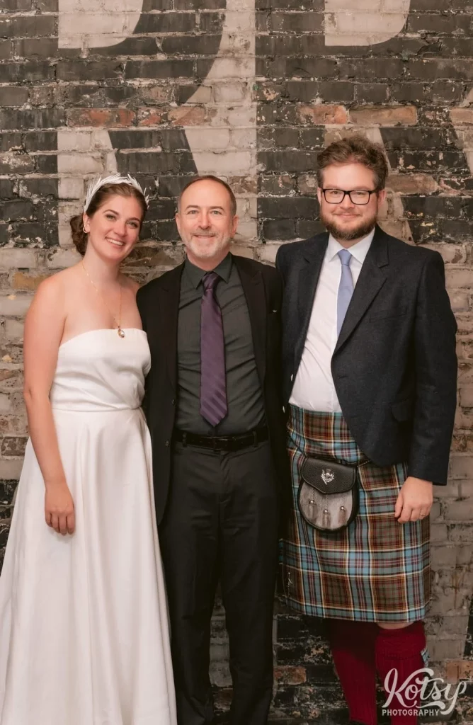 A bride in a white wedding gown and groom in a kilt posed for a photo with her father wearing a black blazer and purple tie during their Burroughes Building wedding reception in Toronto, Canada