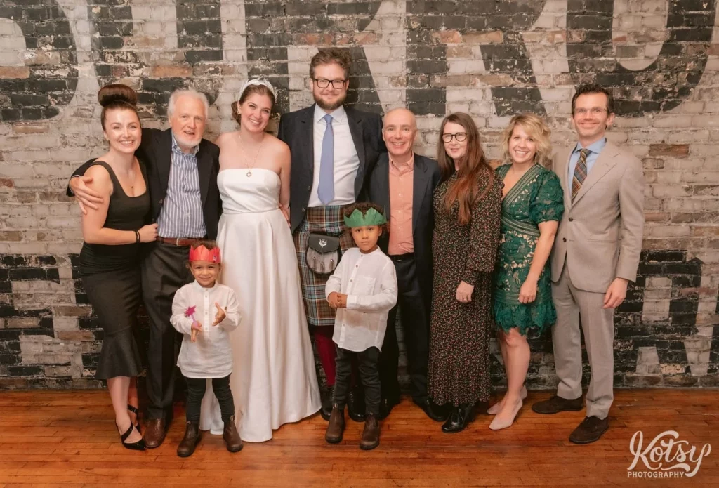 A bright and groom posed for a group photo with some family and children in front of a gray brick wall with black letters during a Burroughes Building wedding reception in Toronto, Canada
