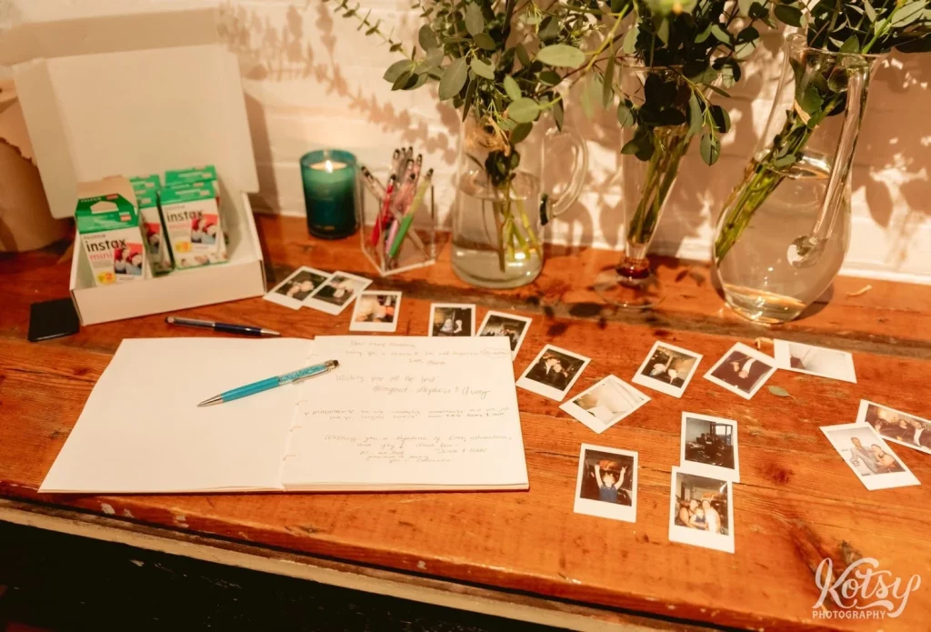 A wooden table with Polaroid Pictures and guestbook as well as dried plants and vases it seemed during Burroughes Building wedding reception in Toronto, Canada