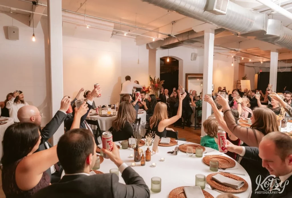 A room full of people sitting at dinner tables raise their glass in cheers during a Burroughes Building wedding reception in Toronto, Canada